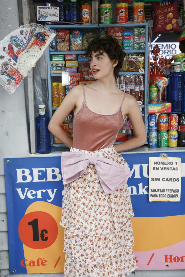 Mujer con falda midi floral Sister Ophelia, de volantes ligeros, lazo lavanda y tejido con brillo de lúrex, posando frente a un kiosko colorido