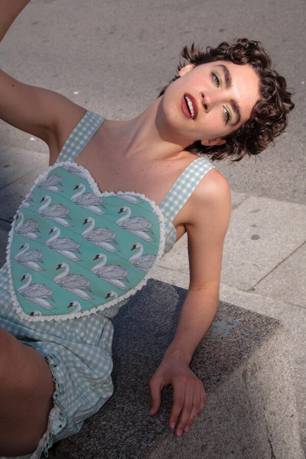 Modelo con top de tirantes setilo corazón en verde agua con estampado de cisnes y cuadros vichy, sonriendo al sol en una calle urbana