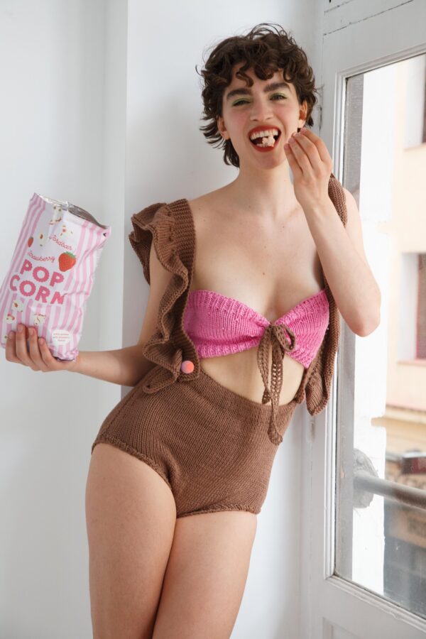 Persona sonriendo, vestida con el conjunto Annette tejido a mano: top rosa bandeau con lazo y short marrón de tiro alto, sosteniendo una bolsa de palomitas de sabor fresa