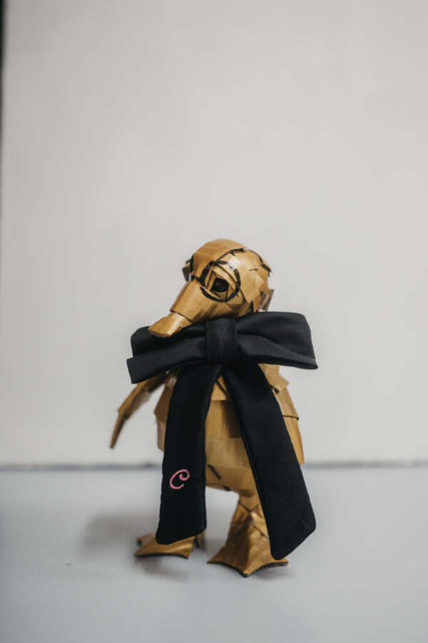 Escultura de cartón en forma de pato dorado con gafas y lazo negro con inicial