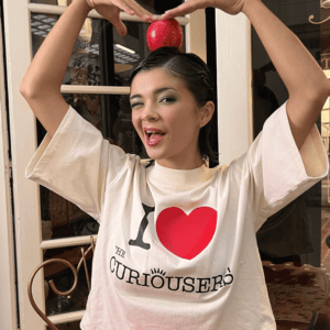 Modelo sujetando manzana en la cabeza y guiñando un ojo con camiseta I ❤️ The Curiousers